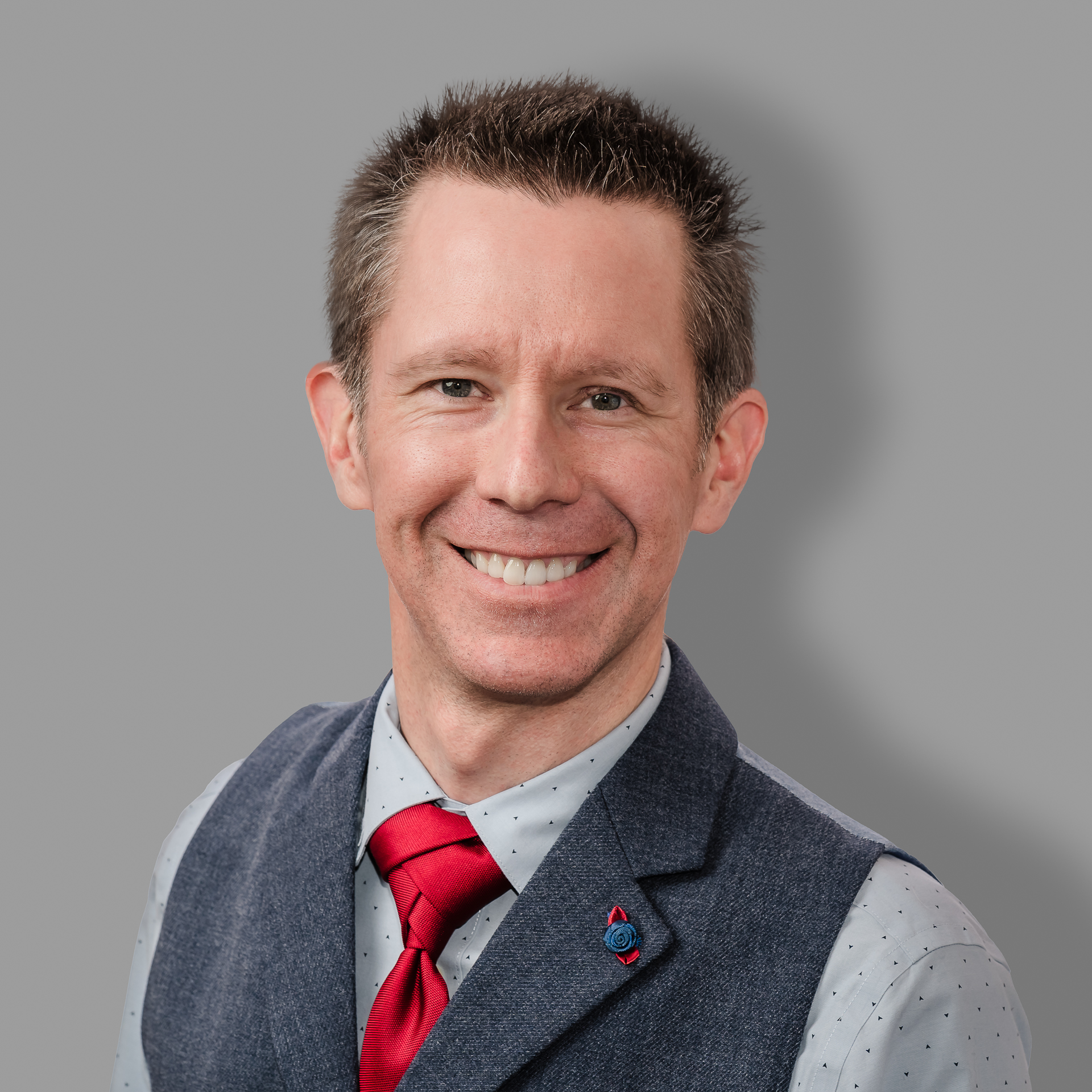 Shane, co-chair of the Education Community of Practice, smiling, wearing a grey shirt, dark grey vest, and a red tie.