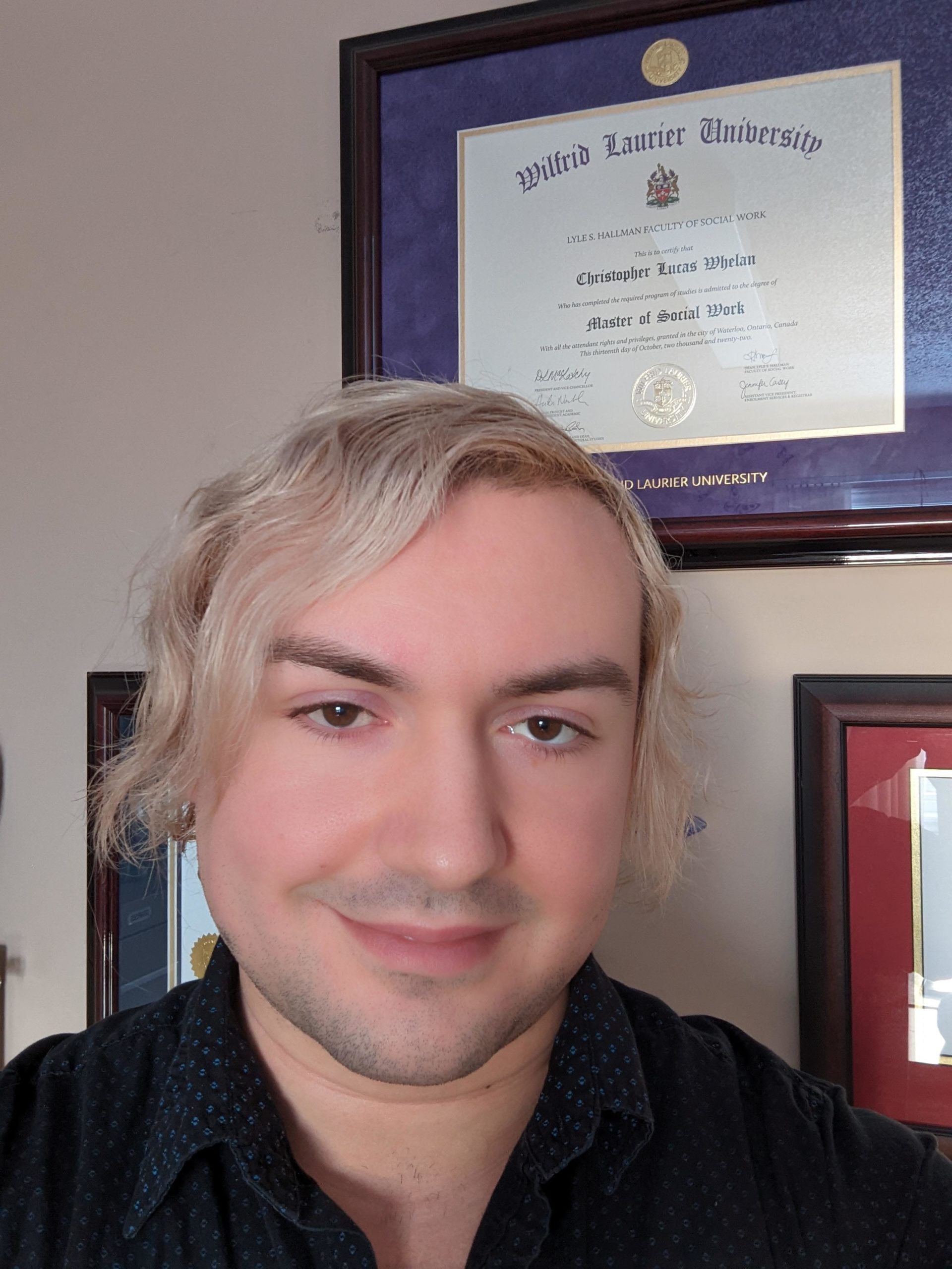 Christopher standing in front of their framed university degrees with blond hair.