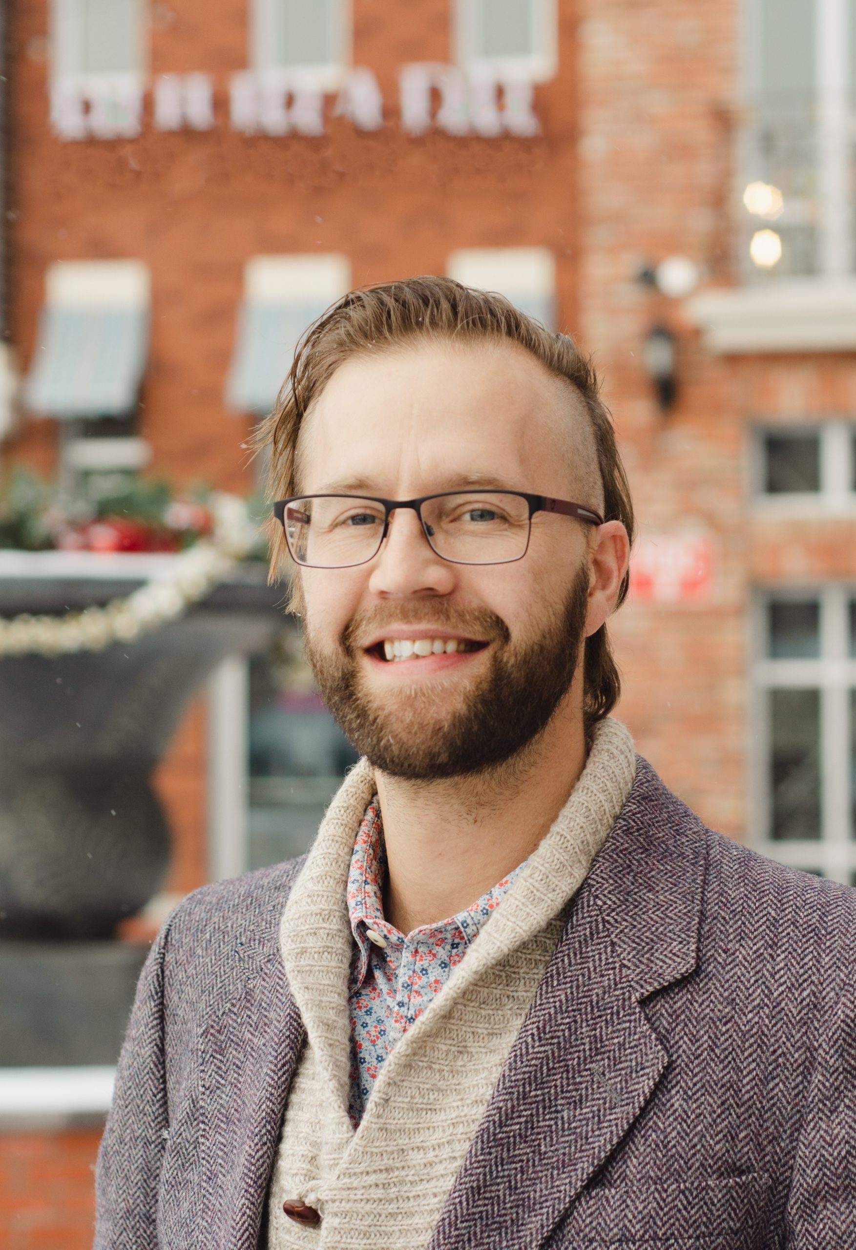 Drewe standing outside wearing glasses and a layered shirt, sweater and wool blazer