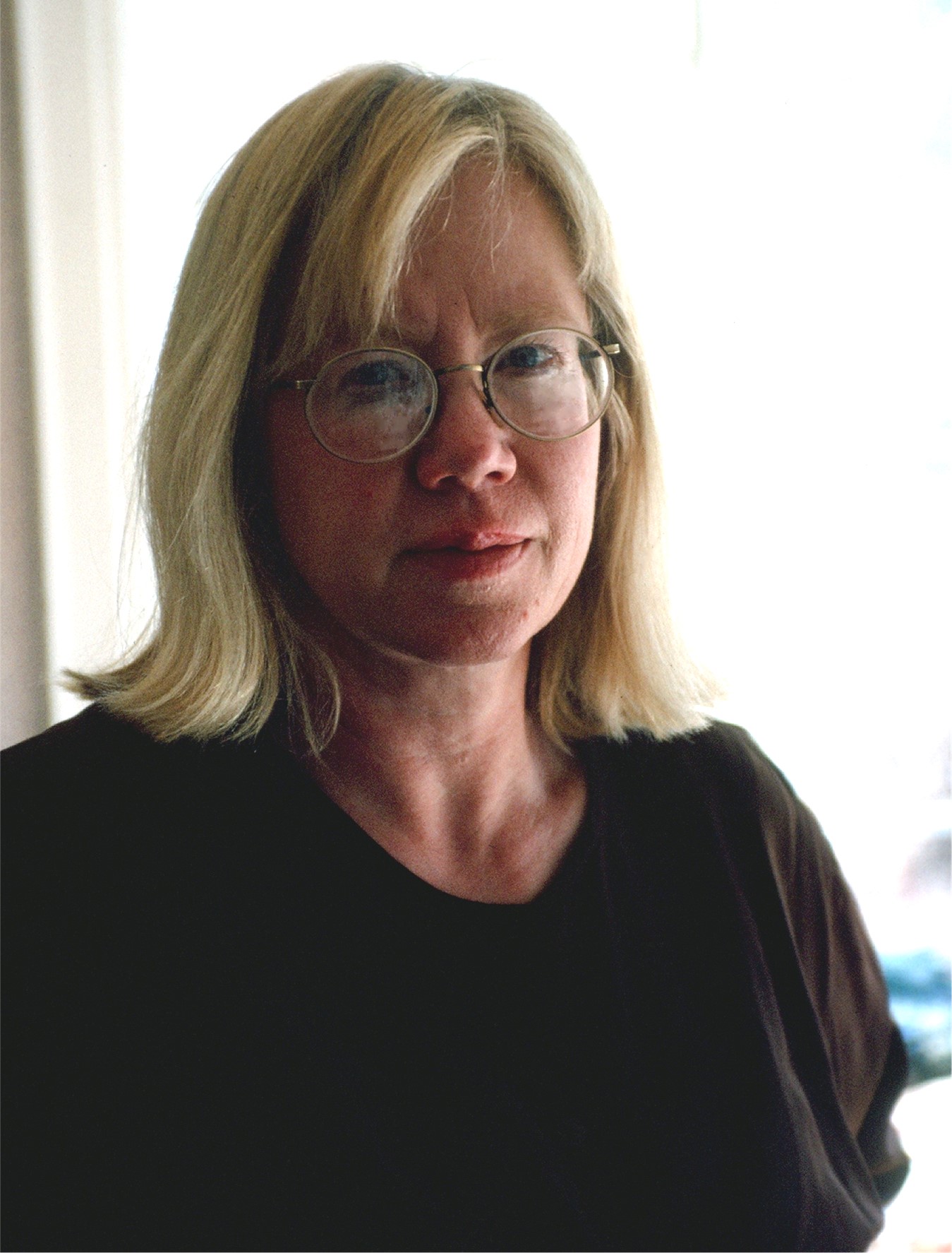 Barbara stands infront of a lighted background. She has blond hair, glasses and is wearing a dark shirt.